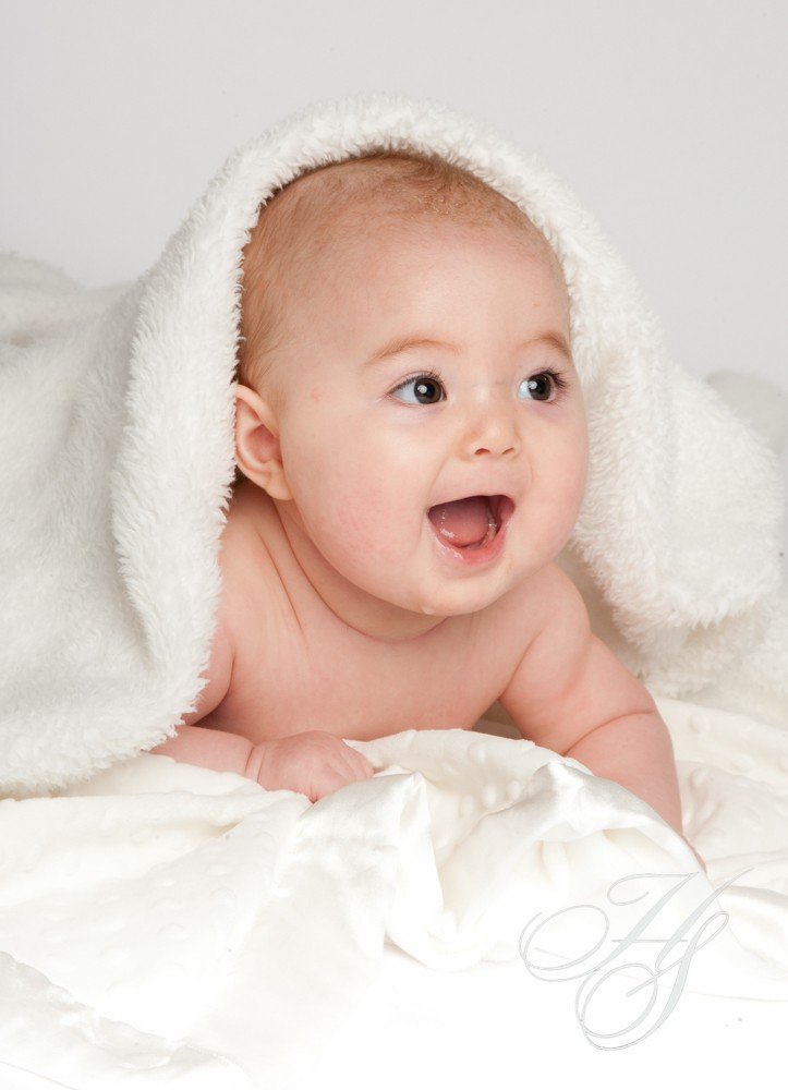 A baby is laying on the bed with his head wrapped in a blanket.