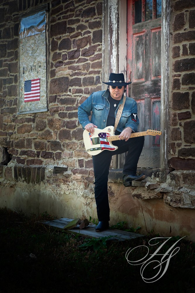 A man in blue jacket playing guitar near brick wall.