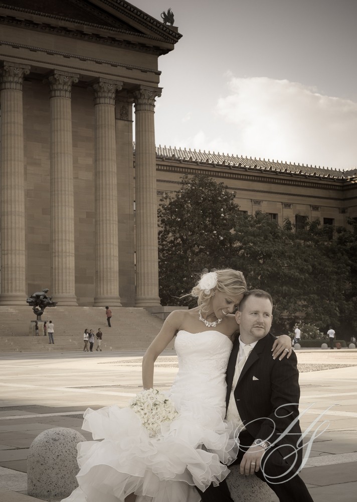 A bride and groom pose for the camera in front of a large building.