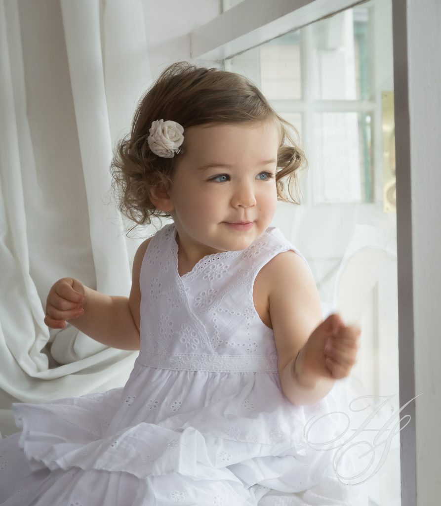 A little girl in white dress standing next to window.